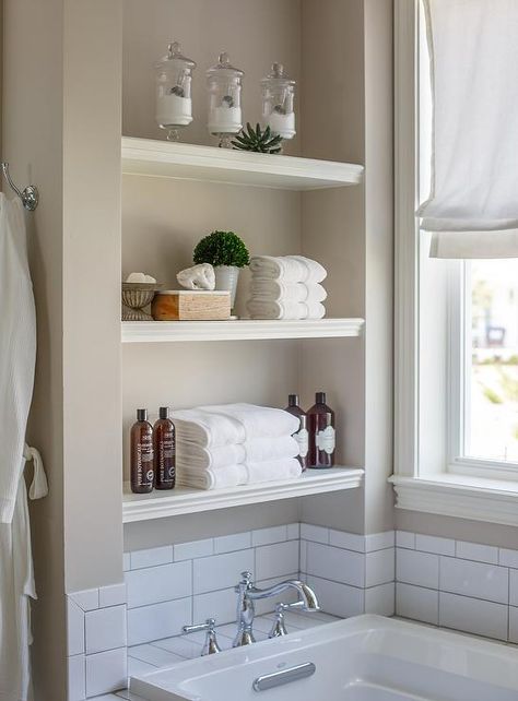 White stacked floating shelves are mounted in a nook above a drop in bathtub fitted with a white subway tiled deck holding a polished nickel vintage hook and spout tub filler in front of white subway backsplash tiles fixed beneath a window dressed in a white roman shade accenting gray wall paint. Bathroom Shelves Above Tub, Bathroom Shelves Over Tub Modern, Shelving Above Tub, Over Tub Shelves, Shelving Over Bathtub, Shelves Above Free Standing Bathtub, Shelf Above Tub, Shelves Above Tub, Bathtub Inset Shelf