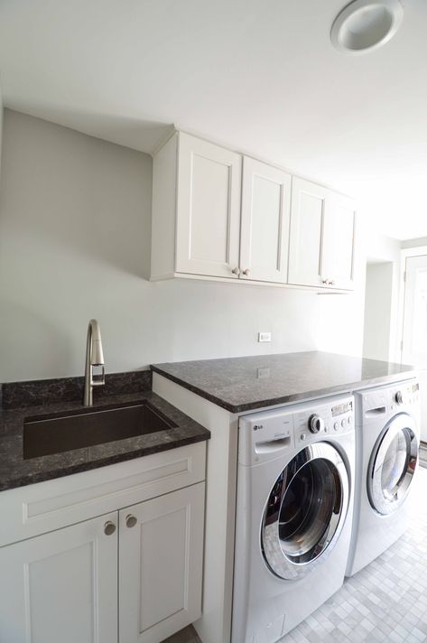 This simple, sleek laundry room has plenty of contrast with dark countertops and white cabinets. The countertops create a perfect folding zone. Soapstone Countertops Laundry Room, Black Countertop Laundry Room, Black Quartz Countertops Laundry Room, Laundry Room Black Countertops, Laundry Room With Fridge, Hinged Countertop Laundry, Laundry Room White Cabinets Black Countertop, Laundry Room Granite Countertop, Black Counter White Cabinets