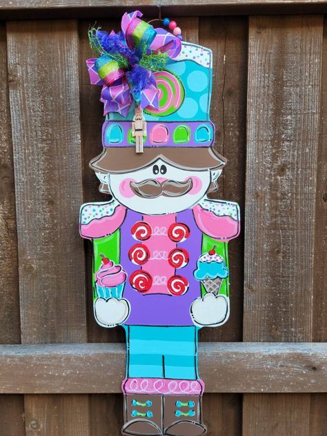 "This darling nutcracker door hanger is ready to greet your Christmas guests at your front door! A nutcracker in shades of pink, purple, blue and green has a sweet tooth!  He has gum drops on his hat and is holding ice cream and cupcakes.  His shoulders and hat are dripping with glittered \"frosting.\" A festive bow adds color and cheer to the bottom of the hand painted door hanger. This wooden door hanger is around 26-28 inches tall.  Custom sizes are available.  Just ask! Also, please let us k Front Door Decor Winter, Gingerbread Nutcracker, Nutcracker Pink, Christmas Front Door Decor, Nutcracker Crafts, Winter Door Hanger, Hanger Christmas Tree, Pink Nutcracker, Door Hanger Christmas