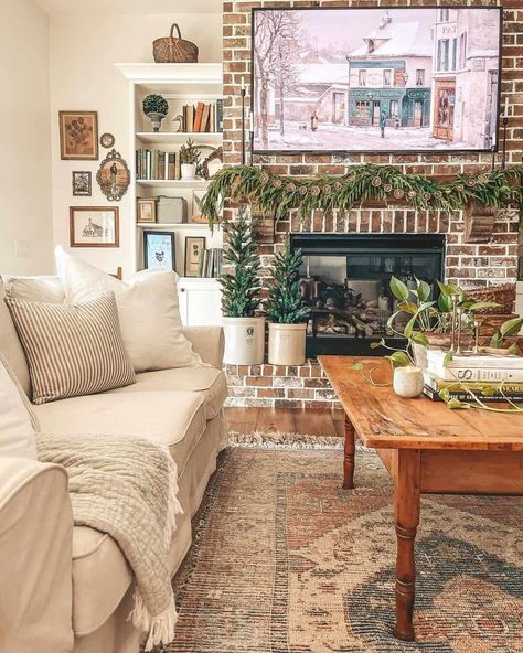 A Pine garland is hung across a red brick fireplace in this living room. Two miniature pine trees in white planters are set on the hearth below, next to a white shelf filled with books. A wood coffee table is decorated with a trailing plant and a votive candle on top of a stack of books...   Image: lakefrontfarmhouse Red Brick Fireplace, Shiplap Living Room, Farmhouse Fireplace Ideas, White Stone Fireplaces, Red Brick Fireplaces, White Brick Fireplace, Wood Coffee Table Rustic, White Mantel, White Shiplap Wall