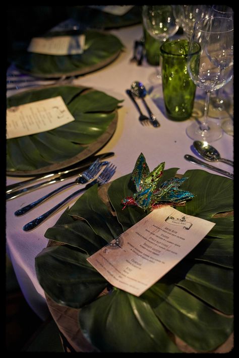 Crossing a second wooden bridge over a flowing river, guests came to a clearing that featured a stunning waterfall. Just on from here guests found their tables dressed with bespoke made elephant print charger plates, banana leaf menu cards, and green glassware. Each table had a palm tree growing through the centre, with tropical birds placed on the branches. Overhead between the trees aerial artists dressed in animal print costumes could be seen performing mesmerising routines throughout. Rainforest Themed Wedding, Rainforest Theme Quince, Rainforest Theme Birthday Party, Rainforest Quinceanera Theme, Safari Prom Theme, Rainforest Theme Wedding, Rainforest Centerpieces, Rainforest Wedding Theme, Garden Of Eden Party Theme