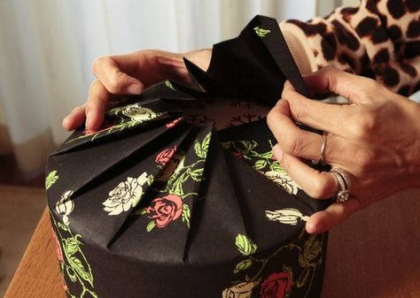 Instead of finding a rectangular box for a round package like a cookie tin, try the fashion trick of pleating. Warning to the easily flustered: This takes practice. For a round cookie tin, Sumika used  Cream & Red Roses on Black  gift wrap, a striking and elegant alternative to traditional reds and greens.       VIDEO: Wrapping round presents      She cut a piece as wide as the circumference of the circle (plus a half-inch extra) and as long as the diameter of the lid plus one-half the height o… Japanese Gift Wrapping, Gift Wrapping Techniques, Wrapping Presents, Japanese Gifts, How To Wrap, Gift Wraps, Gift Wrap Ideas, Creative Gift Wrapping, Diy Gift Wrapping