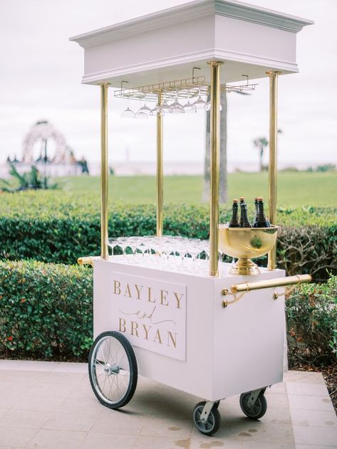 A white wedding bar cart with gold accents, featuring a champagne holder, glass display, and a personalized sign. Bourbon Bar Wedding, Wedding Bar Setup, Wedding Bar Cart, Outdoor Wedding Bar, Champagne Cart, Wedding Bar Decor, Wedding Drink Station, Bar Setup, Indoor Wedding Receptions