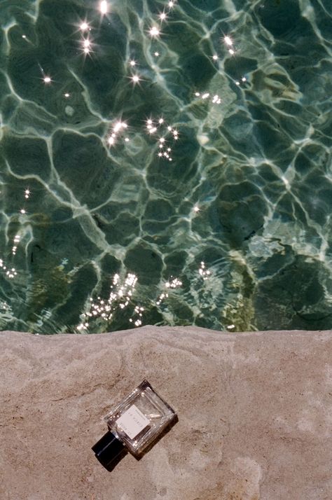 a perfume bottle lies on a rock next to a blue-green body of water that is shimmering in the sun light . All Natural Perfume, Vetiver Oil, Sandalwood Oil, Cedarwood Oil, Patchouli Oil, Bergamot Oil, Rose Fragrance, Natural Perfume, Clean Skincare