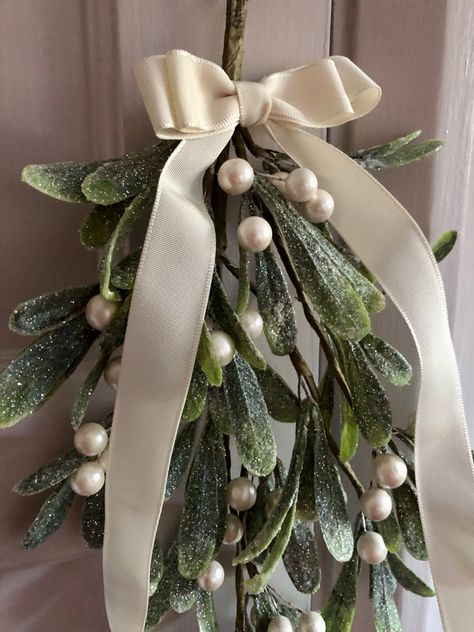 ‘Christmas Kisses’ Traditionally hung above doorways at Christmastime, it’s said that kissing under the mistletoe brings good luck for the year ahead. Handmade from a generous bunch of quality faux mistletoe, dusted with pretty white fine glitter and adorned with pearlised berries. Tied with a fabric bow in your choice of classic ivory or festive red. Complete with integral hook. Average length of 40cms. Mistletoe Decoration Christmas, Mistletoe At Wedding, Mistletoe Wedding Decoration, Mistletoe Centerpiece, Mistletoe Doorway, Christmas Mistletoe Aesthetic, Bow Christmas Decor, Mistletoe Ideas, Mistletoe Aesthetic