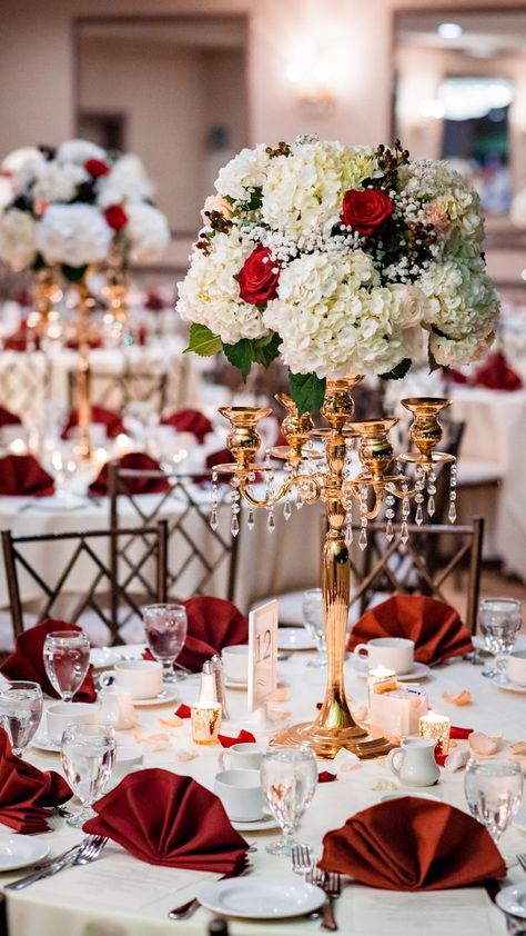 Romantic red and ivory floral wedding centerpieces on bronze metallic stand with hanging acrylic beads. Classic white tablecloth with luxury red napkins decorated with red rose petals and glassware. Red Gold White Wedding Theme, Champagne And Red Wedding Decorations, Ivory Red And Gold Wedding, Wedding Red Gold Theme, Wedding Decor Red And Gold, Red White And Gold Wedding Theme, Wine Gold And White Wedding Decor, White And Bronze Wedding Decor, Red And White Wedding Theme Decor