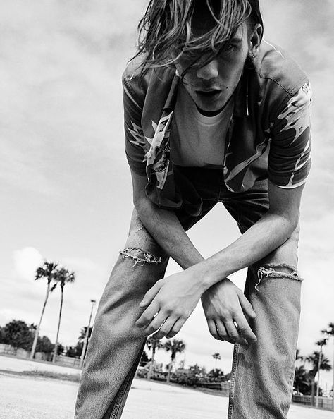 The Road Trip Part 2 - Black and White image of a guy bending over and looking straight down at the camera. He is wearing a short sleeve Hawaiian shirt, open over a white t-shirt and denim jeans with splits on the knees. The Kooks, Jackets Summer, White Image, 2024 Collection, Creepers, Bending, Summer Essentials, Leather Jackets, White T Shirt