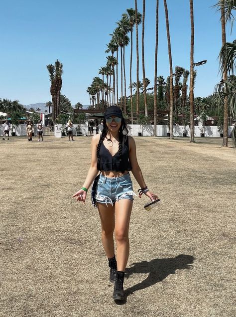 Young woman with black hair in 2 braids, black Alo hat, black v-neck crop tank, ripped denim shorts, blue reflective sunglasses, and black combat boots; she is walking towards the camera with a shaka in one hand and empty cup in the other, she is smiling with teeth and mouth slightly open; a line of palm trees runs diagonally in the background; the sky is blue and clear Jean Shorts Outfit Festival, Taco Festival Outfit, Denim Shorts Festival Outfit, Festival Outfits Midsize, Casual Coachella Outfits, Denim Shorts Festival, Summer Festival Outfit Ideas, Trendy Festival Jean Shorts, Denim Festival Shorts