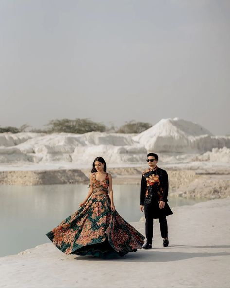 The bride-to-be is dressed in an elegant, flowy lehenga with vibrant floral prints, which brings a sense of movement and grace to the image. Her long, wavy hair cascades over her shoulders as she walks, adding a touch of soft femininity to the composition. The groom-to-be complements her look in a black kurta with matching floral accents, exuding a contemporary yet traditional vibe. The composition of the shot feels balanced and artistic, with the groom standing relaxed against the tree whil... Jaipur Prewedding Shoot, Jaipur Pre Wedding Shoot, Jaipur Prewedding, Flowy Lehenga, Prewedding Shoot, Black Kurta, Pre Wedding Poses, Photo Board, Pre Wedding Shoot