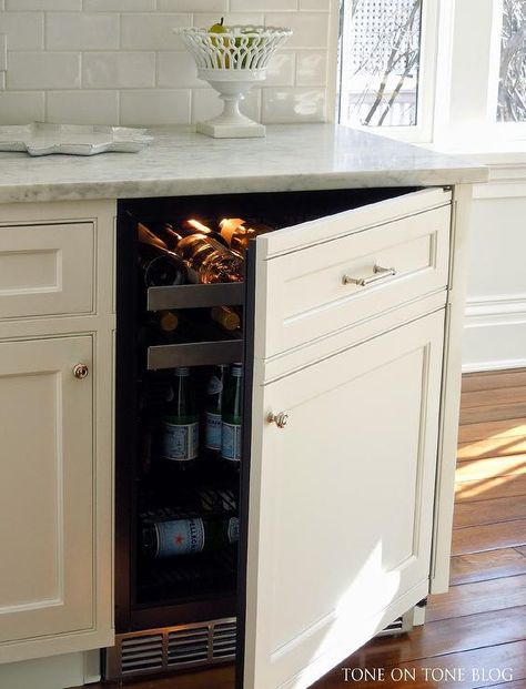 Kitchen features creamy white cabinets fitted with a white paneled beverage fridge with pull out wine trays paired with carrera marble countertops and a glazed white subway tiled backsplash. Inside Cabinet Wine Storage, Hidden Wine Fridge Cabinet, Under Counter Wine Fridge In Kitchen, Hidden Drink Fridge, Paneled Mini Fridge, Corner Wine Fridge, Wine Fridge Inside Cabinet, Beverage Refrigerator Cabinet, Fridge That Blends In With Cabinets