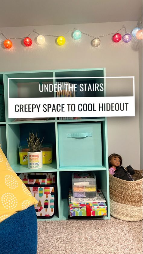 An aqua colored bookshelf sits in an under the stairs area with colored string lights and a basket of kid toys. Under The Stairs Toy Storage, Understairs Kids Space, Under The Stairs Library, Under Stairs Play Area Small Spaces, Under Stairs Closet Playroom, Closet Under Stairs Ideas For Kids, Under Stairs Toy Storage, Under Stairs Ideas For Kids, Under Stairs Kids Space