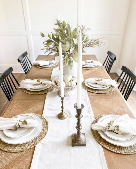 A white board and batten accent wall is displayed at the end of a wooden dining table. A white runner with elegant decorations stretched the length of the table, including round, rattan placemats, and white plates. Black spindle back chairs are placed in front of table settings, with a large leafy bouquet placed in a white jug in the center of the table. Home Table Decorations, Dining Table Decor With Runner, Daily Dining Table Setting, Placemats And Table Runners, Table Setting Wooden Table, Neutral Dining Table Setting, Table With Placemats, Wood Dining Table Place Setting, Dining Table With Placemats