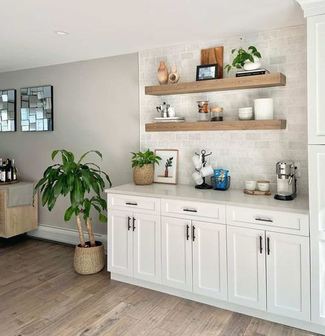 Two long floating shelves hang on an offset off-white tiled kitchen wall. A stainless steel coffee maker, a black stand holding white mugs, and a plant in a wicker basket are placed on the light grey counter on white cabinets with black pulls. A large plant in a wicker basket is placed on the warm wood floor next to the white cabinets. Tiled Kitchen Wall, Open Kitchen Shelves Ideas, Blue Dining Room Walls, Light Wood Dining Table, Black Pulls, Stainless Steel Coffee Maker, Warm Wood Flooring, Kitchen Floating Shelves, Long Floating Shelves