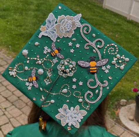 Aesthetic green emerald honey bee themed simple elegant graduation cap decoration with flowers and rhinestones, pearls and bee decals that is sparkly Green Graduation Aesthetic, Graduation Cap Ideas Simple, Elegant Grad Cap Ideas, Grad Cap Inspo High School, Graduation Cap Simple Design, Bee Graduation Cap, Flower Graduation Cap Designs, Grad Caps Aesthetic, Pretty Grad Caps