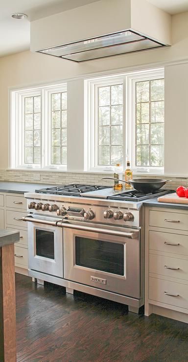 Lovely kitchen features ivory shaker cabinets paired with Pietra Cardosa Limestone countertops and a white and gray  linear tiled backsplash placed under windows and a ceiling mount vent hood. Oven Hood In Front Of Window, Kitchen Stove Under Window, Oven Under Window In Kitchen, Stove Top In Front Of Window, Stovetop In Front Of Window, Oven In Front Of Kitchen Window, Oven By Window, Range In Front Of Window Kitchen, Range With Window Behind