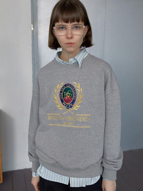 The Classic Sweatshirt depicted here offers a blend of traditional collegiate style and modern casual wear. It features a distinctive crest graphic on the front, with elaborate details and text that reads BRENDA BRENDEN EST. 1980, adding a touch of preppy flair. The gray hue of the sweatshirt makes it a versatile piece that can be easily paired with various outfits, while its crew neck and ribbed cuffs ensure a comfortable and snug fit. - The crest graphic is richly designed, making the sweatshirt stand out as a statement piece while still retaining a classic look.- Made from soft, durable fabric, this sweatshirt provides warmth and comfort, ideal for cooler days.- Its traditional fit and simple color allow it to be styled effortlessly with both casual jeans and more formal trousers.- Collegiate Sweatshirt Outfit, Sweat Shirt Outfits Women, Graphic Crewneck Outfit, Collared Shirt Under Sweatshirt Outfit, Shirt Under Sweatshirt, Grey Sweatshirt Outfit, Crew Neck Sweatshirt Outfit, College Sweatshirt Outfit, Gray Sweatshirt Outfit