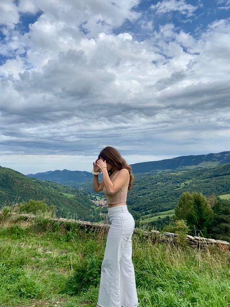 Young girl in nature looking down with hands on face. Mountains in the backgrownd. Brown long hair, beige corset top, lilac cardigan and white flared pants. All green nature and cludy, moody sky. Mountain Photo Poses, Hill Photoshoot Ideas, Forest Waterfall Aesthetic, Mountain Outfit Aesthetic, Aesthetic Photos Nature, Hill Station Outfit Ideas, Mountain Photography People, Mountain Photo Ideas Instagram, Corset Top Outfit Ideas