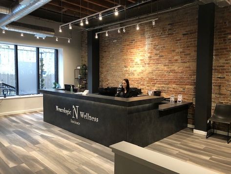 This black desk really stands out against the light wooden floor. Exposed brick long with the open ceiling and track lights adds an industrial vibe. #design #chiropractic #lobby Brick Interior Office, Office Exposed Brick, Industrial Chic Lobby, Exposed Brick Office Space, Front Desk Office Design, Industrial Clinic Design, Office Wooden Flooring, Brick Office Design, Lobby Industrial Design