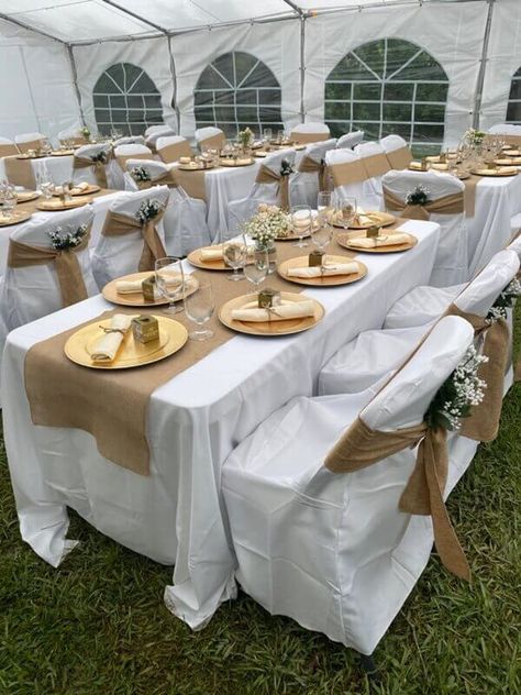 Exude rustic vibes with your outdoor wedding décor setup by dressing up your tables with white tablecloths and burlap runners. Also, cover your chairs with white chair covers along with burlap chair sashes and tuck in white baby breath florals for an ethereal touch. Wow your guests with an impressive place setting using gold chargers, burlap napkins, and favor boxes. Moreover, add gorgeous visual appeal to your arrangement by arranging alluring florals in glass vases as your table centerpieces. Brown And White Centerpieces, White Table Cloth With Gold Runner, Rustic Wedding Table Set Up, Chair Covers Wedding Reception, Outdoor Events Decor, Outdoor Night Wedding, Burlap Chair Sashes, Burlap Centerpieces, Bautizo Ideas