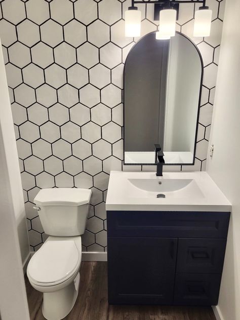 A bathroom wall is lined with white hexagon tiles and black grout. A white toilet is on the left side and a white sink atop a dark dresser is on the right. Black accents on the light fixtures, modern taps in sink and accents on arched mirror atop the sink pop with the dark grout. Hexagon Tiles, Tile Stores, Honeycomb Pattern, Endless Possibilities, Wall Tiles, Honeycomb, Tile, Bee, Square