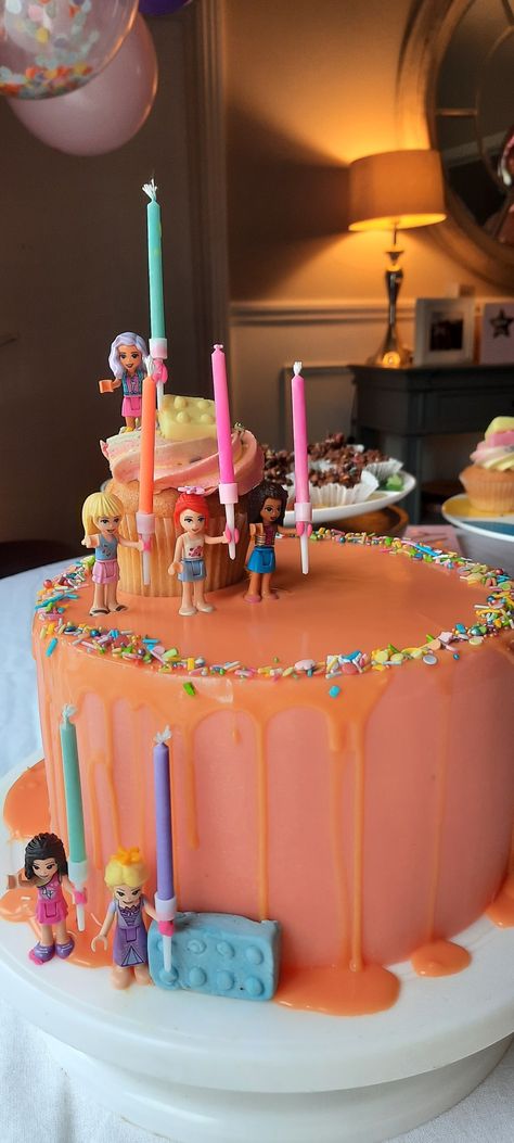 Three tier iced buttercream cake, pink frosting and orange white chocolate gnache drip with lego girls holding multicolored birthday candles! Lego Princess Cake, Lego Themed Birthday Party Girl, Lego Friends Birthday Party Ideas, Lego Birthday Party Girl, Lego Birthday Cake Girl, Lego Friends Birthday Cake, Birthday Cake 7th Girl, Lego Friends Party Ideas, Girl Lego Birthday Party