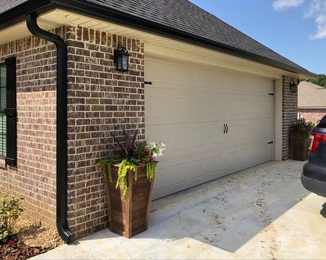 Added black gutters and magnetic hardware to the gatage door. Built the tall planters and stainedto match the front doors and columns ❤️ Brown Brick Black Shutters, Black Gutters Tan House, Black Gutters Red Brick House, Black Gutters Gray House, Tan House Exterior, Garage Colors, Black Gutters, Magnetic Hardware, Outside Paint Colors