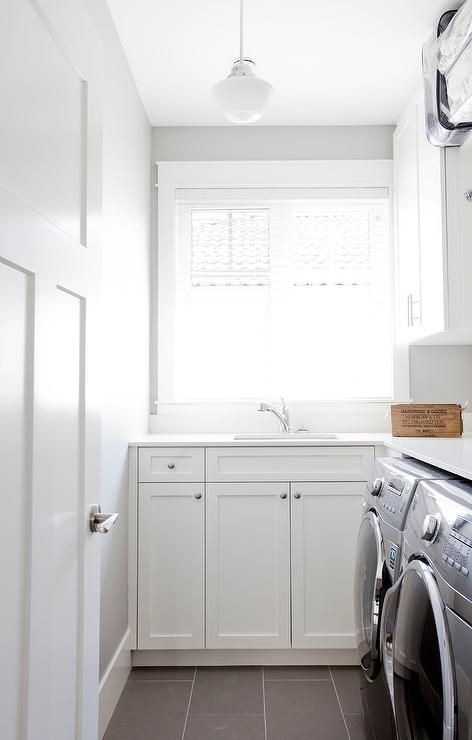 Clad in gray slate floor tiles, this well design small white mudroom is fitted with a gray front loading washer and dryer enclosed beneath an l-shaped white countertop fitted with a sink and a polished nickel faucet positioned beneath a window framed by gray walls and located next to white shaker upper cabinets lit by a school house pendant. L Shaped Utility Room Small Laundry, Small L Shaped Laundry Room, L Shape Laundry Room Layout, L Shape Laundry Room, L Shape Laundry, L Shaped Laundry Room Layout, L Shaped Laundry, L Shaped Laundry Room, White Mudroom