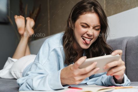 Image of woman showing her tongue and playing online game on cellphone by vadymvdrobot. Image of woman showing her tongue and playing online game on cellphone while lying on sofa at home #Sponsored #tongue, #playing, #online, #Image Playing On Phone Pose, Person Playing Video Games Reference, Playing Video Games Pose Reference, Holding Phone Reference, Working With Laptop, Gesture Drawing Poses, Woman Smiling, Doing Homework, Chair Pose