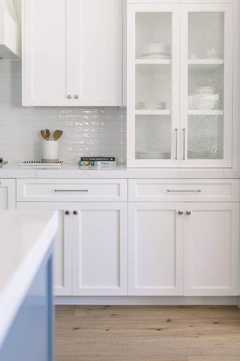 White shaker kitchen cabinets accented with chrome hardware and a white quartz countertop are fixed beneath seeded glass cabinets and against white glazed backsplash tiles. Seeded Glass Cabinets, Kitchen Hardware Trends, White Shaker Kitchen Cabinets, Glass Kitchen Cabinets, White Shaker Kitchen, White Shaker Cabinets, Shaker Kitchen Cabinets, Kitchen Knobs, White Quartz Countertop