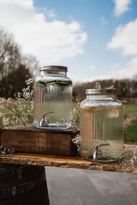 This image shows a drinks station at a wedding. The focus is on two glass dispenser containers, with cucumber and lemon water in them. One of the containers is displayed on a crate, with dried flowers surrounding the bar. Double Drink Dispenser, Drink Dispensers Wedding, Drink Dispenser Stand Diy, Drink Display Wedding, Bakery Theme, Drink Dispenser Stand, Diy Pedestal, Welcome Drinks, Hobbit Party