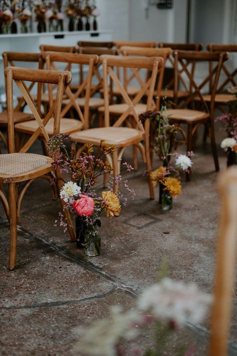 Small floral arrangements in bright colours with retro vibe sit beside wooden chairs lining the aisle | Wedding Aisle Centerpieces, Affordable Wedding Aisle Decorations, Wild Flower Wedding Chairs, Top Of Aisle Decor, Inexpensive Aisle Decorations Wedding, Simple Isle Decoration, Minimal Isle Decor, End Of Aisle Arrangements, Wedding Aisle Planters