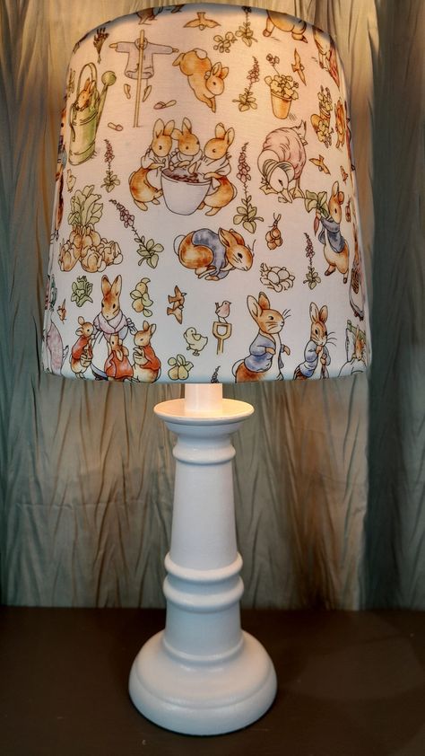 This sweet accent / Nursery lamp features a shade upholstered in a lovely Beatrix Potter pattern depicting Peter Rabbit and his family. The shade features a front and back with two finished off side seams. Please refer to the photo gallery as well as the video. The shade fits on any lamp base, sitting on the socket above the light bulb on a suspended ring. The shade itself measures 6 and 3/4 in High by 7 and 1/2 in wide and fits a base up to 12 in height. The shade can be purchased alone or with the lamp base. The assembled lamp offered is 15 in tall. The hand painted and sealed base comes in your choice of white or light gray. Base color options can be viewed in the photo gallery. What a delightful gift this would make for any new baby or for any small child who loves the Fairy Tale of Pe Peter Rabbit Nursery Sculptures & Statues, Beatrix Potter Bedroom, Story Book Nursery Theme, Beatrix Potter Nursery Gender Neutral, Beatrice Potter Nursery, Beatrix Potter Nursery Ideas, Peter Rabbit Nursery Ideas, Nursery Ideas Whimsical, Vintage Peter Rabbit Nursery