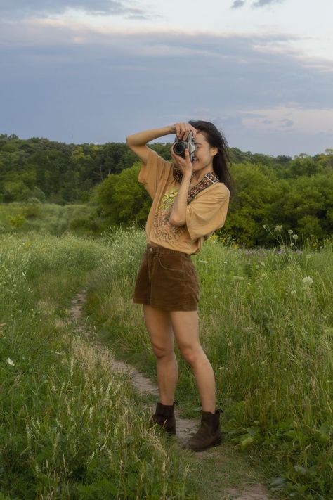 A girl standing in a field taking a photo with a film camera. She is wearing a tan graphic tee, brown corduroy shorts, and brown boots. Earth Tone Outfits Women Summer, Summer Outfits Earth Tones, Earth Tones Summer Outfits, Loose Earthy Outfits, Earth Tones Wardrobe, Earthy Going Out Outfit, Granola Lesbian Style, Casual Earth Tone Outfits, Earthy Tone Clothes