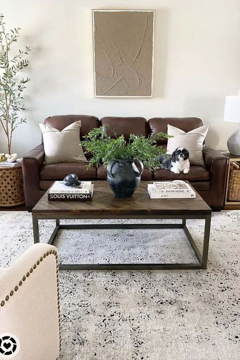 Step into a cozy haven with this living room featuring modern plaster artwork in warm brown tones. The artwork graces a comfortable brown leather couch. Beside the couch, you’ll find wooden farmhouse side tables, one of which cleverly features an open bottom for convenient storage in a woven basket. A herringbone-patterned wooden coffee table adds a touch of elegance, adorned with lush greenery in a black jug, all resting gracefully on an antique area rug. Farmhouse Side Tables, Light Wood Furniture, Sheepskin Blanket, Plaster Artwork, Brown Leather Couch Living Room, Modern Farmhouse Living Room Ideas, Mocha Cream, Authentic Farmhouse, Soft Autumn Color Palette