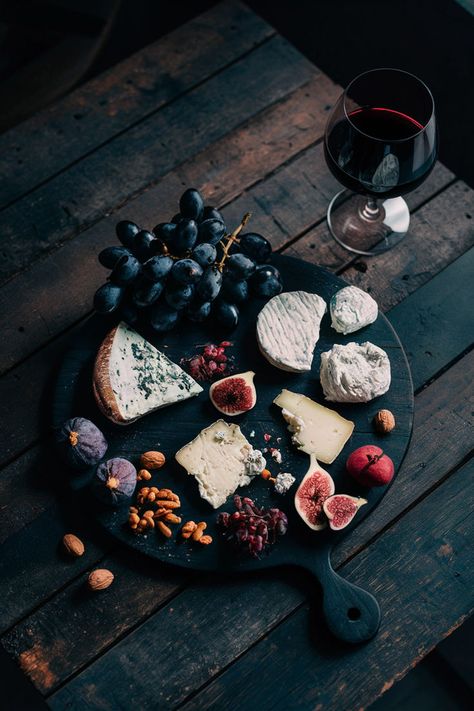 Dark wooden cheese board with assorted cheeses, figs, black grapes, nuts, and a glass of red wine, set on a rustic wooden table with moody lighting. Dark Charcuterie Board Ideas, Charcuterie Board Photoshoot, Minimal Charcuterie Board, Gothic Charcuterie Table, Moody Charcuterie Board, Cheeseboard Aesthetic, Dark Charcuterie Board, Charcuterie Aesthetic, Black Charcuterie Board