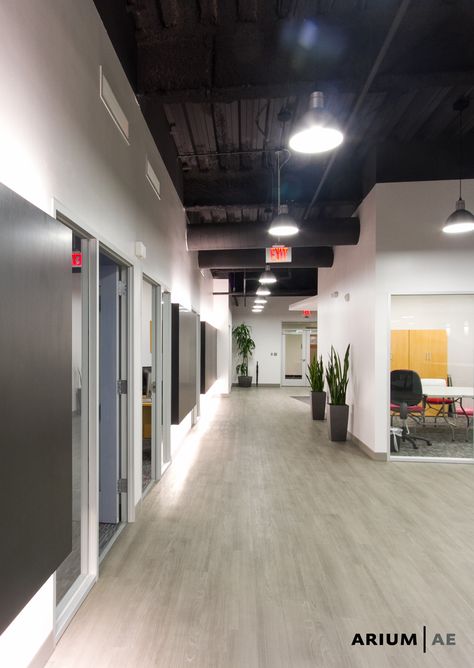 Corridor in an office space, with black laminate accent wall with light coves, exposed ceiling painted black, vinyl wood-look laminate floors Black Ceiling Office Design, Exposed Office Ceiling, Black Ceiling Office Interiors, Office Exposed Ceiling Design, Office With Black Ceiling, Exposed Ceiling Office Design, Dark Office Ceiling, Black Office Ceiling, Industrial Exposed Ceiling