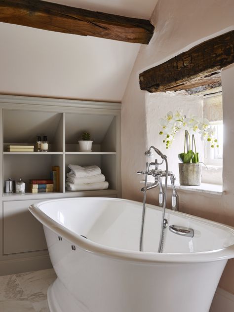 Sloping ceilings and heavy beams can present design challenges in older properties. The bathroom featured was designed to maximise storage with cabinetry and shelving handmade to fit the available space under the eaves. Freestanding bath from @vandabaths, taps and accessories from @samuel_heath and furniture finished in #purbeckstone from @farrowball.⁠ ⁠ #bathrooms #ensuite ⁠ #bathroom #bathroomdesign ⁠ #bathroomcabinetry #bath ⁠ #bathroomdesigners ⁠ #bespokebathroom⁠ #yorkshirebathroom⁠ Bathroom Eaves Storage, Bathroom Sloping Ceiling, Eaves Bathroom Storage, Bathroom In The Eaves, Shower Under Sloped Ceiling, Bathrooms With Sloping Ceilings, Exposed Beams Bathroom, Luxury Cottage Bathroom, Bathroom With Eaves