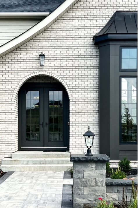 This brick arched entryway is such a dreamy way to enter this stunning home! The creamy white exterior and light stonework are contrasted by the black doors, windows, roof and lighting to create a breathtaking exterior aesthetic!  New Construction | Home Trends 2024 | Home Design | Minnesota Home Trends | Black & White Aesthetic White Brick House Black Trim, Elegant Home Exterior, Arched Entryway, Exterior Aesthetic, Arch Entryway, Farmhouse Garage, Build Design, Brick Arch, Exterior Inspiration