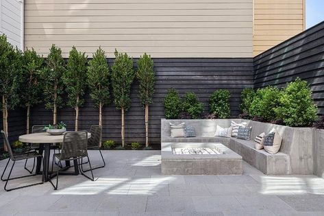 A built-in l-shaped concrete sofa sits in a backyard at a concrete firepit, while gray rope chairs surround a concrete and metal dining table positioned on concrete pavers. Large Concrete Pavers, Round Concrete Dining Table, Paver Fire Pit, Concrete Patio Designs, Rectangular Fire Pit, Contemporary Patio, Concrete Bench, Concrete Fire Pits, Concrete Dining Table