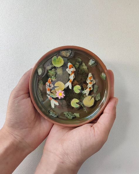 From dessert to decor.🎏⛩️ . I thoroughly enjoyed putting this koi fish pond together. Sometimes it's good to take a break from what you usually do to see what more you can get done. I repurposed a clay bowl to create this mini koi fish pond using acrylic-painted sand, natural stones, grass, and resin layers. After laying down the base and adding a few stones, I submerged tiny handmade clay koi fish in resin and topped it off with delicate lotus leaves and a lotus flower. Love how this came to... Clay Koi Pond, Resin Koi Pond, Clay Koi Fish, Clay Pond, Clay Fish, Clay Things, Koi Fish Pond, Clay Bowl, Lotus Leaves