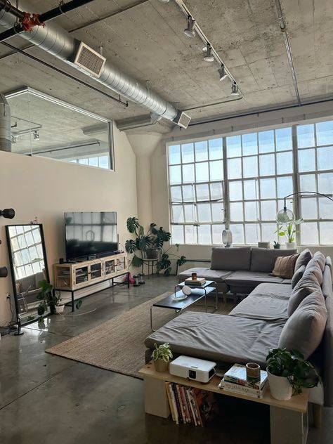 This stylish industrial loft features an open layout with large windows, concrete ceilings, and exposed ductwork. The cozy sectional sofa, minimalist decor, and natural elements like plants and a woven rug create a warm and inviting living space. Perfect inspiration for modern urban living! Urban Loft Apartment Industrial Style, Exposed Ductwork Ceiling, New York Loft Apartment Industrial, Vintage Loft Apartment, Industrial Studio Apartment, Industrial Cozy, Industrial Loft Living Room, Nyc Loft Apartment, Cozy Industrial Living Room