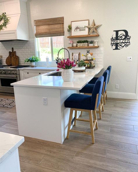 Tall blue chairs sit on a grey wood floor behind a white kitchen peninsula. Floating wood shelves on a light gray wall hang above a rustic white tile backsplash. A stainless steel stove sits beneath a white hood. Kitchen With Sink In Peninsula, One Wall Kitchen With Peninsula, Peninsula Wall Ideas, Sink In Peninsula Kitchen, Kitchen Ideas With Peninsula, Farmhouse Kitchen Peninsula, Kitchen Peninsula With Sink, Small Kitchen With Peninsula Layout, Medium Kitchen Ideas