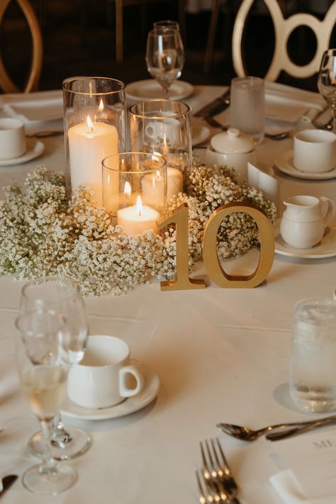 Cylinder vase wedding centrepiece surrounded by babies breath and gold table number sign Wedding Table Decorations Round Tables Candles, Cylinder Vase Centerpiece Wedding Floating Candles White Flowers, White And Gold Table Centerpieces, Simple Gold Wedding Centerpieces, White And Gold Centerpiece, Floating Candles And Babys Breath, Candles Centrepiece Wedding, Babys Breath And Candles Centerpiece, Baby's Breath Wedding Table Decor