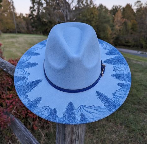 This hand burnt wide brim fedora makes a perfect addition to any wardrobe. The faux suede baby blue material burns a gorgeous shade of dark navy blue allowing the design to stand out. Each mountain is intricately burned by hand with a pyrography machine using various tips to achieve depth and a rock-like texture. Pine trees are burnt in front of the mountains creating a beautiful landscape. The design is original and completely freehanded. For a finishing touch the hat is adorned with a dark blu Hat Burn Designs, Cowboy Hat Crafts, Burned Hats, Hat Painting, Cowboy Hat Design, Hat Burning, Mountain Hat, Country Hats, Cowgirl Gifts