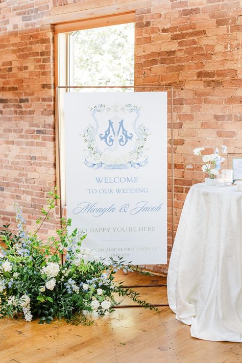 a custom wedding monogram printed on a hanging welcome sign for a wedding beside a welcome cocktail table with a white patterned linen on it and a grounded floral arrangement beside it Wedding Crest Welcome Sign, Wedding Welcome Sign With Monogram, Cocktail Hour Sign, Welcome Table Wedding, Stradley Davidson, Wedding Welcome Table, Wedding Cocktail Hour, Blue And White Wedding, Cocktail Hour Wedding