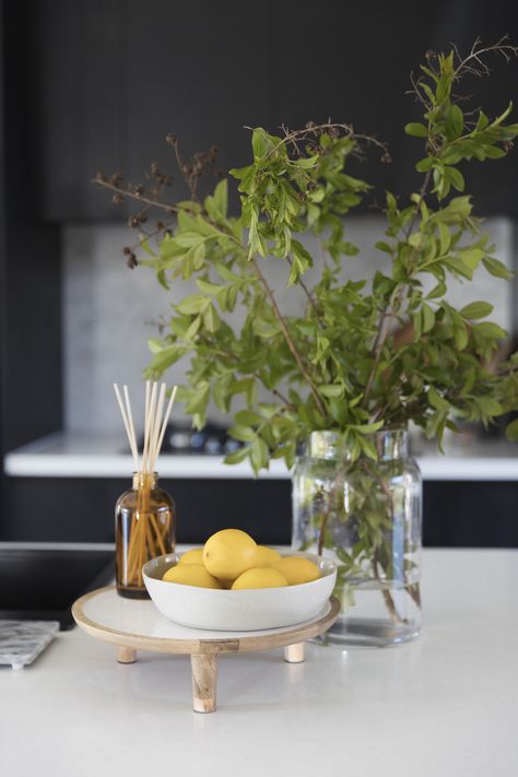 Vignettes or styled arrangements add interest to surface areas of your home. They are also a great way to display your favourite items! You can put them on your coffee tables, sideboards, kitchen benches, beside table, shelves and so much more! We give you the best tips and tricks on how to create and style a vignette in your space. Kitchen vigentte styling with stylised tray, aroma diffuser, bowl of fruits, lemons, large vase, foraged branches. Click the pin to see more vignette inspo and tips! Kitchen Bench Fruit Bowl, Fruit Bowl Decor Centerpieces, Fruit Bowl On Island, Farmhouse Fruit Bowl, Bowl Of Lemons Decor, Lemon Bowl Decor, Wooden Risers Display Kitchen, How To Display Fruit On Counter, Fruit Display Ideas Kitchen