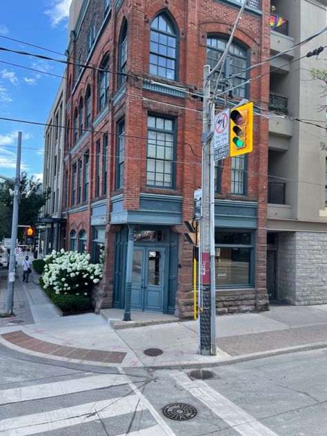 An old red-brick, corner building in urban Toronto. You are looking at the corner and can see two full stories of this house. The windows are blue and very tall, the door is at the corner of the house and is blue as well. Going down the left side are white beds of flowers in front of the building. 
You can also see a street light which is green and a part of the street. Life In Usa, Manifesting 2024, Toronto Street, Old Toronto, Toronto Photos, City Scapes, Toronto City, Street Corner, O Canada