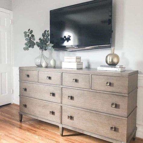 A large eight-drawer dresser with an ashy wood-toned finish is placed beneath a large flatscreen TV in a modern farmhouse bedroom. Add some square brass hardware for a metallic texture to go with the golden glass vase along the top of the dresser...   Image: thisblissfulhome Tv Above Dresser Bedroom, Bedroom Dresser Decor With Tv, Tall Floor Mirror, Dresser Decor With Tv Above, White Antique Dresser, Short Dresser, Small White Bedrooms, Grey And Gold Bedroom, White Wood Dresser