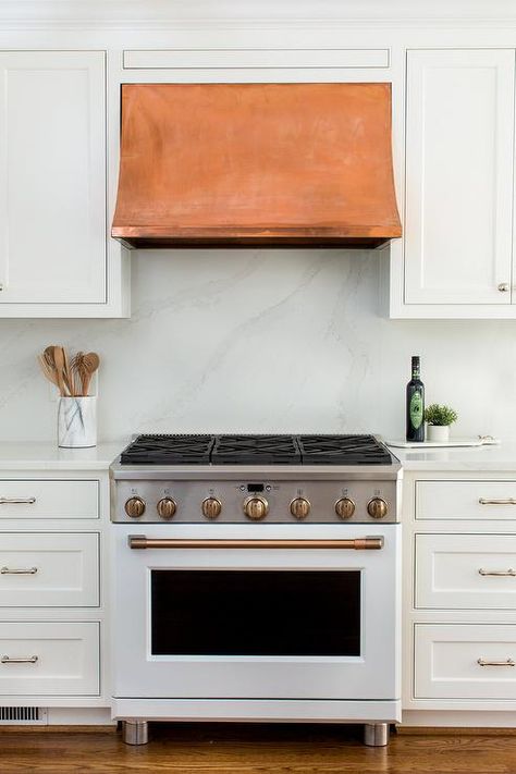 Fixed between white inset cabinets, a copper range hood is mounted against a honed marble backsplash and over a white stove with brass trim flanked by white cabinets topped with a honed marble top. White Kitchen Vent Hood, Small Space Kitchen Ideas, Bm Swiss Coffee, Montgomery Homes, White Stove, Fish Shack, Blue Kitchen Island, Kitchen Island Tops, Inset Cabinetry