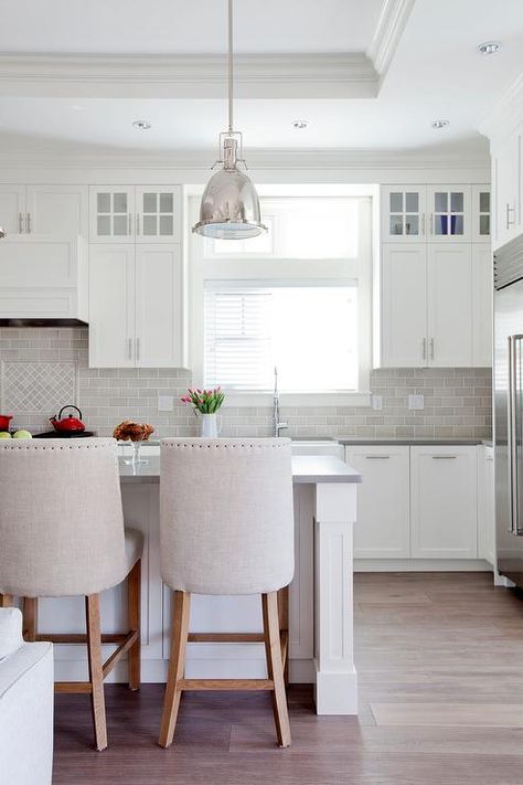 A stainless steel fridge is recessed beneath cabinets in a wall adjacent to window flanked by white shaker cabinets with nickel pulls located beneath glass front display cabinets and  positioned above a wide farmhouse sink with a pull out polished nickel faucet placed against light gray mini subway backsplash tiles as a white kitchen hood is mounted above diamond pattern cooktop tiles. White Kitchen Hood, Kitchen White Cabinets, Backsplash For White Cabinets, Backsplash With White Cabinets, Trendy Kitchen Tile, Kitchen Natural, Nickel Faucet, Trendy Kitchen Backsplash, Subway Backsplash